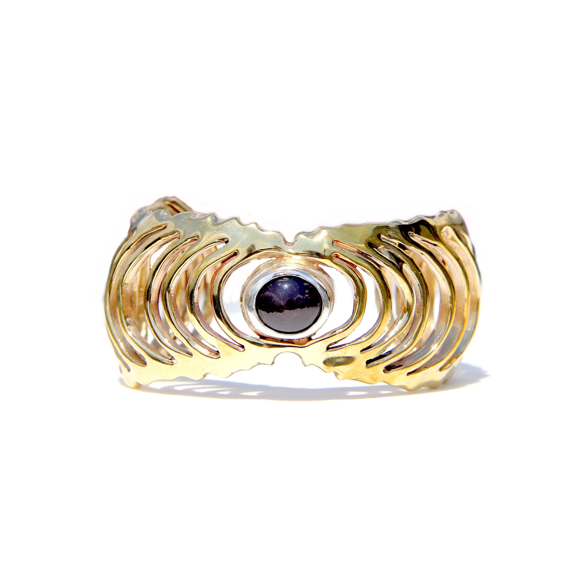 A gold-toned textured cuff  featuring a central oval dark purple/red gemstone, set against a plain white background.